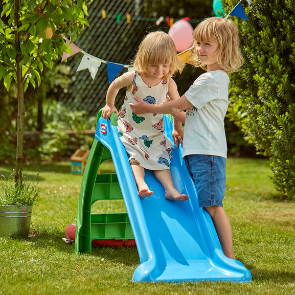 Two children playing on a blue slide in a garden setting with greenery and balloons.