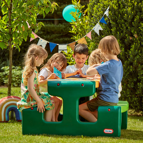 Children playing at a green outdoor picnic table in a garden setting.