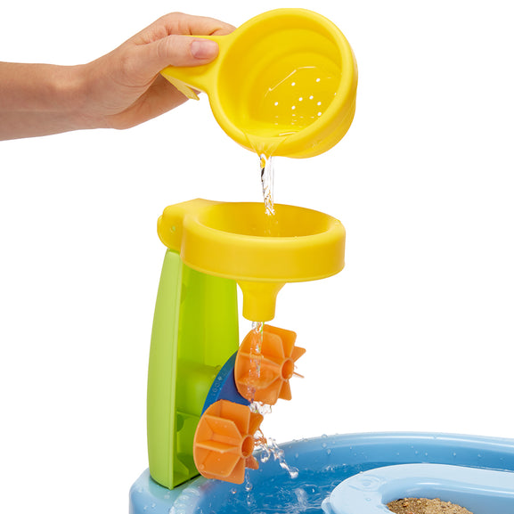 Child's hand pouring water from a yellow funnel into a blue sandpit with a green and orange water feature.