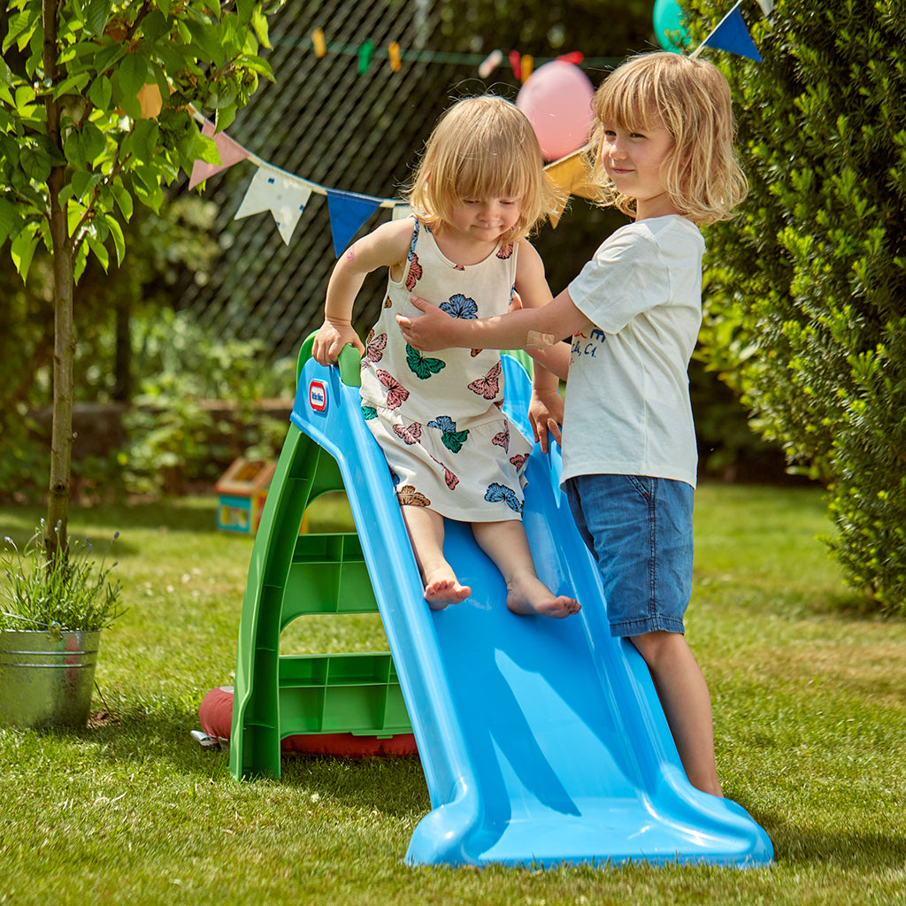 Two children playing on a blue slide in a garden setting with greenery and balloons.
