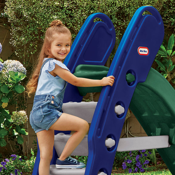 Young girl climbing a blue and green Little Tikes slide with plants and flowers in the background