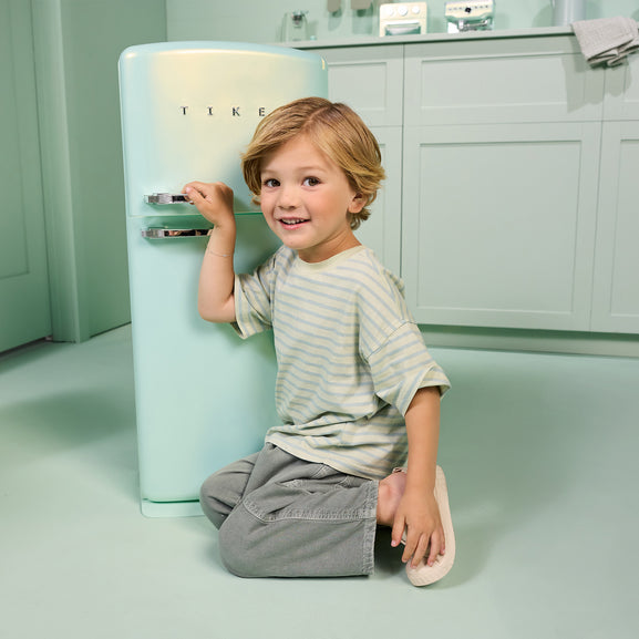 A young child with blonde hair kneels on the floor next to a retro-style mint green refrigerator in a matching pastel kitchen setting. The refrigerator has the brand name 'TIKE' visible on the front.