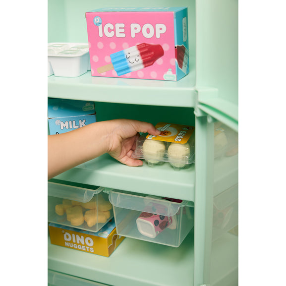 A hand placing a clear plastic drawer organizer into a mint green refrigerator shelf, with other toy food items and a pink 'ICE POP' box visible on the shelves.