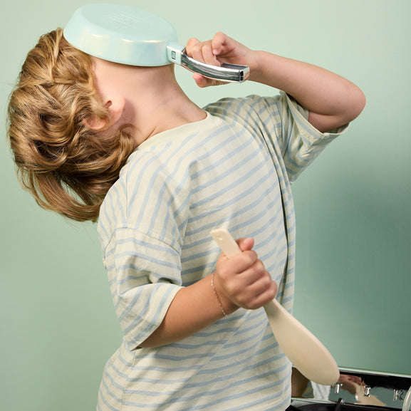 A child wearing a light blue and white striped shirt holds a light blue frying pan against their face while playing with a wooden spoon, set against a solid pale green background.