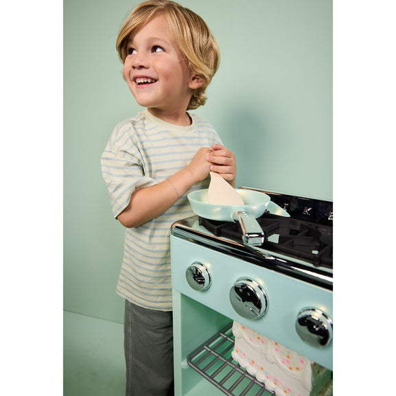 A young child with blonde hair stands next to a mint green toy stove, holding a play pan and spatula. The child is wearing a light beige and white striped t-shirt and grey pants.