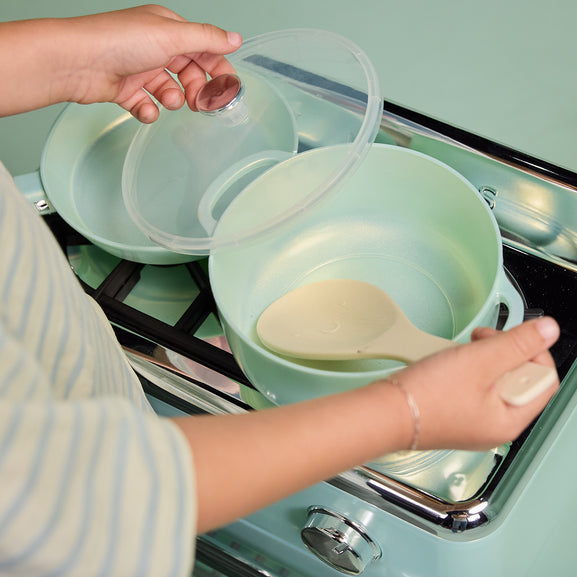 A person's hands are shown handling a mint green cooking pot and its matching lid on a retro-style mint green gas stove. A beige wooden spoon rests inside the pot.