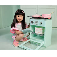 Load image into Gallery viewer, A young girl with long black hair wearing a pink and white dress crouches next to a mint green toy play kitchen stove. She is holding a gray play oven mitt, with the toy stove showing a pink pot, a pink cake, and an open oven door.
