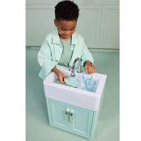 Load image into Gallery viewer, A young child wearing a mint green shirt and matching jacket is playing with a miniature toy kitchen sink and cabinet. The toy sink is white with a mint green base, a chrome faucet, and two clear plastic drinking glasses.
