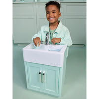 Load image into Gallery viewer, A smiling young boy stands behind a small mint green and white vanity sink, touching the chrome faucet. The scene appears to be a toy or play kitchen setup with matching mint green cabinets in the background.
