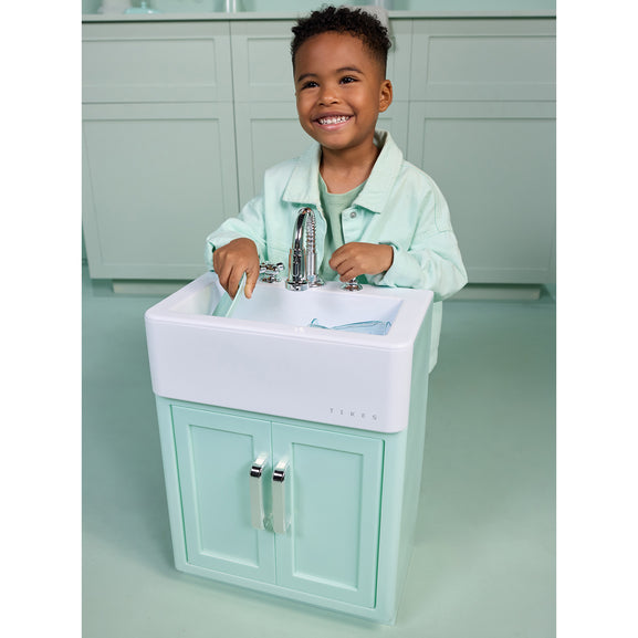 A smiling young boy stands behind a small mint green and white vanity sink, touching the chrome faucet. The scene appears to be a toy or play kitchen setup with matching mint green cabinets in the background.