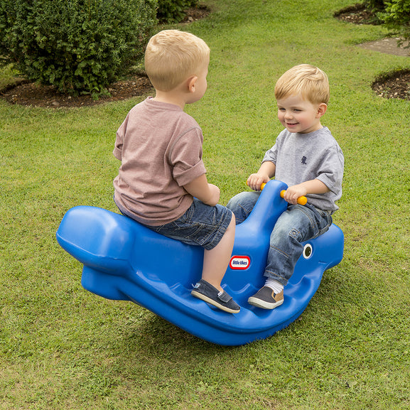 Two children playing on a blue toy rocker in a grassy area.
