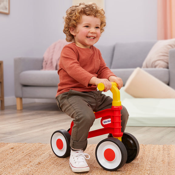 Child sitting on a red and yellow toy tricycle in a living room.