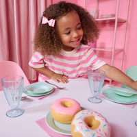 Load image into Gallery viewer, A young girl with curly hair wearing a pink and white striped shirt sits at a pastel pink table setting, looking at two oversized toy donuts in the foreground.

