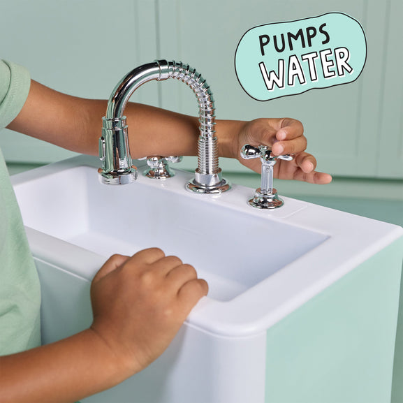 A child's hands and arms are shown interacting with a chrome faucet and handles on a white sink. The background is a mint green paneled wall, and a light blue-green speech bubble in the top right reads 'PUMPS WATER'.