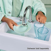 Load image into Gallery viewer, A child in a mint green uniform washes light blue dishes in a white sink under a chrome faucet, with two light blue glasses and a matching plate. Text at the bottom reads 'Dishware Sold Separately'.
