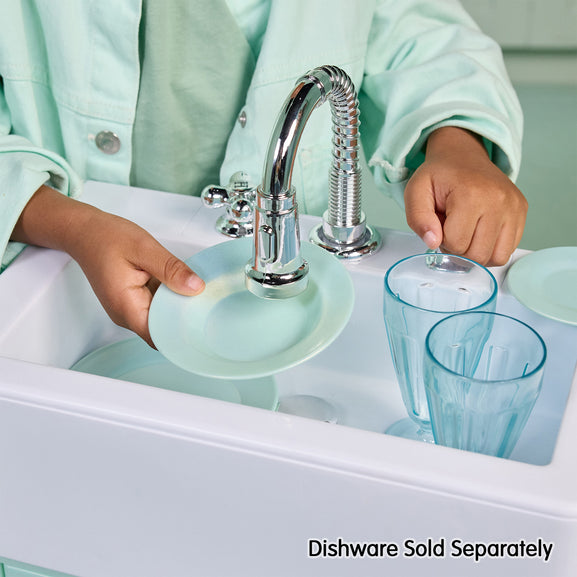 A child in a mint green uniform washes light blue dishes in a white sink under a chrome faucet, with two light blue glasses and a matching plate. Text at the bottom reads 'Dishware Sold Separately'.
